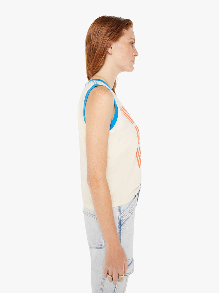 A side view of a woman wearing an off white ringer tank featuring contrast blue trim and a red graphic, paired with a light blue wash jean.