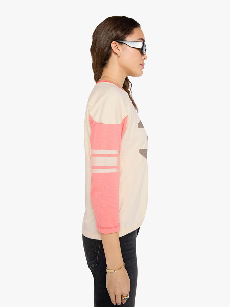 Side view of a woman in off-white with pale pink sleeves and trim, off-white stripes at the elbows, the number 3 in grey on the front and a colorful sewn patch on the hip in a 3/4-length sleeve top with a crew neck, drop shoulders and a boxy fit. 