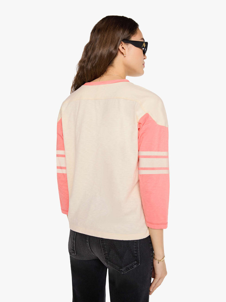Back view of a woman in off-white with pale pink sleeves and trim, off-white stripes at the elbows, the number 3 in grey on the front and a colorful sewn patch on the hip in a 3/4-length sleeve top with a crew neck, drop shoulders and a boxy fit. 
