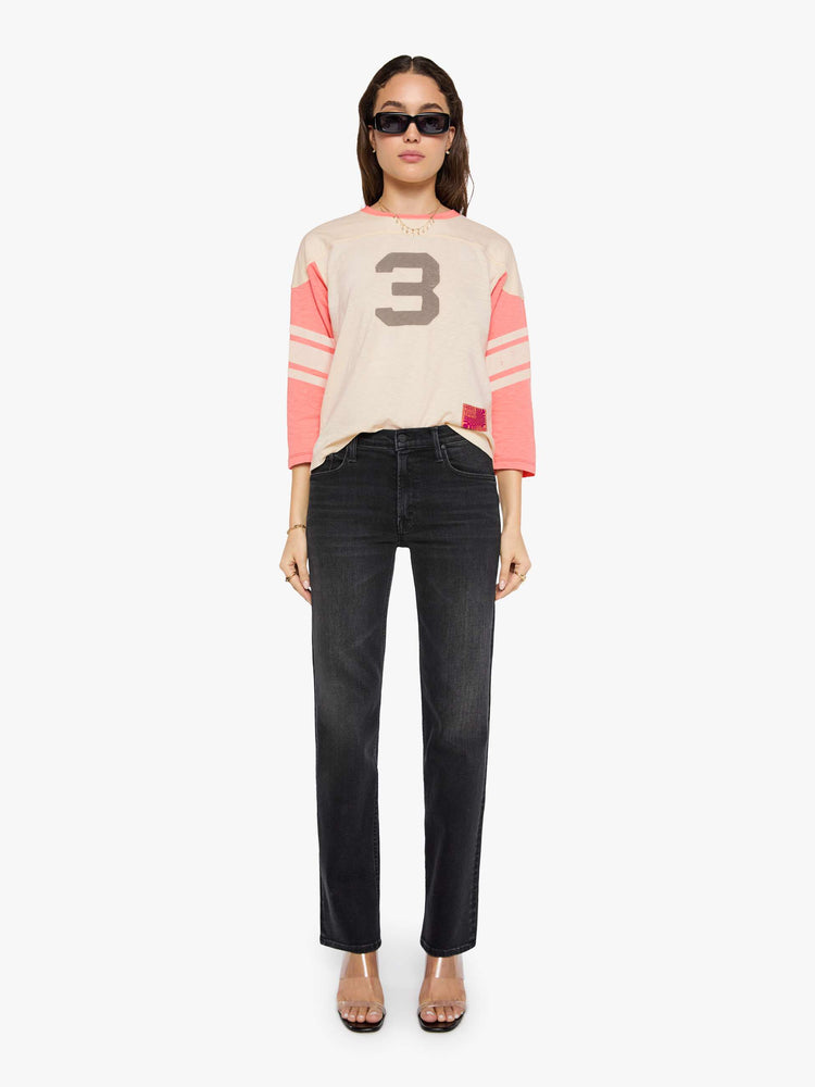Full body view of a woman in off-white with pale pink sleeves and trim, off-white stripes at the elbows, the number 3 in grey on the front and a colorful sewn patch on the hip in a 3/4-length sleeve top with a crew neck, drop shoulders and a boxy fit. 