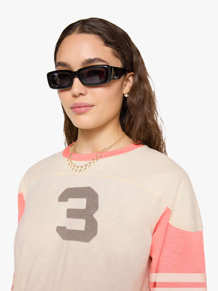 Close up view of a woman in off-white with pale pink sleeves and trim, off-white stripes at the elbows, the number 3 in grey on the front and a colorful sewn patch on the hip in a 3/4-length sleeve top with a crew neck, drop shoulders and a boxy fit. 
