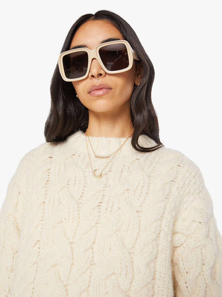 Close up view of a woman in an off white cable crewneck sweater in MOTHERS  stitched in black.