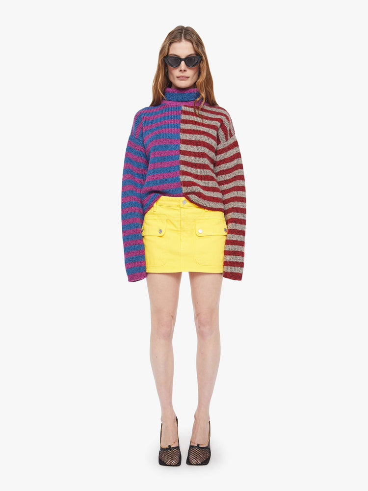 Full body view of a woman in a half blue with purple stripes and half grey with red stripes turtleneck sweater with drop shoulders, extra-long sleeves and a boxy fit.