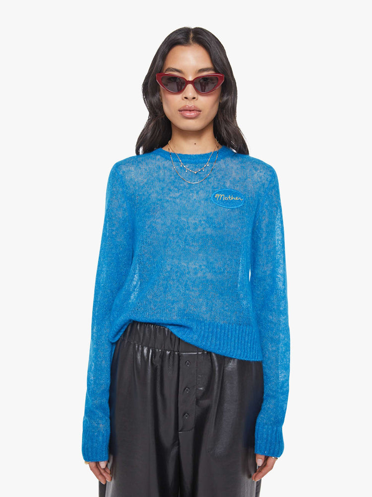 Front view of a woman in a sheer blue with an oval patch on the chest sweater.