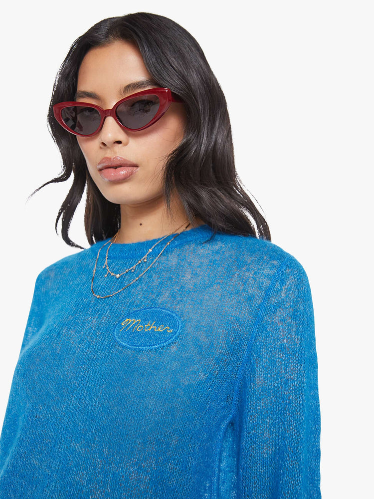 Close up view of a woman in a sheer blue with an oval patch on the chest sweater.