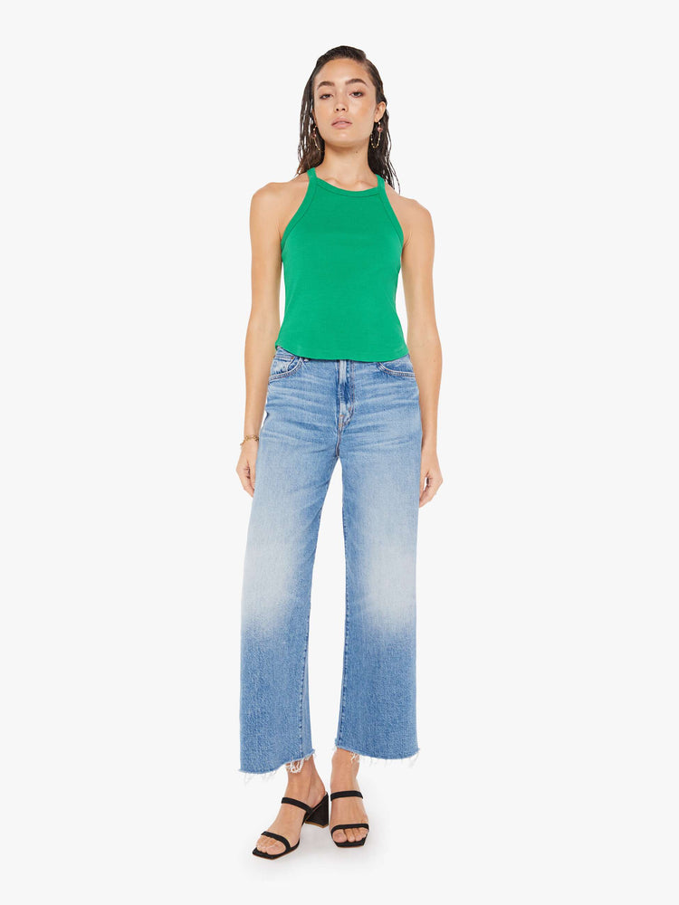 Front full body view of a woman wearing a green ribbed tank top with a cropped curved hem, paired with a medium blue wash wide leg jeans.