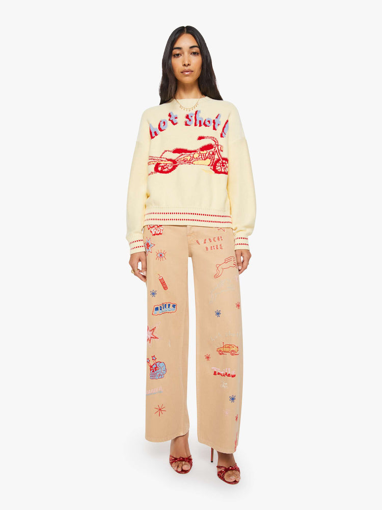 Full body view of a woman in a butter yellow with red and baby blue text, a motorcycle graphic and decorative trim sweater.