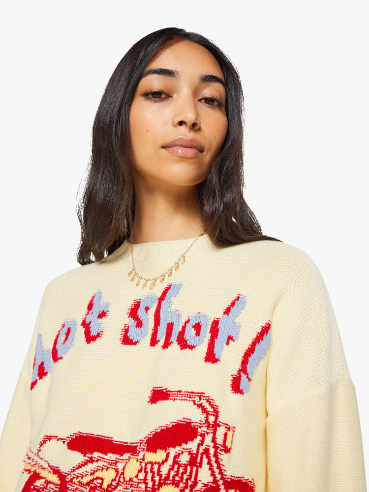 Close up view of a woman in a butter yellow with red and baby blue text, a motorcycle graphic and decorative trim sweater.