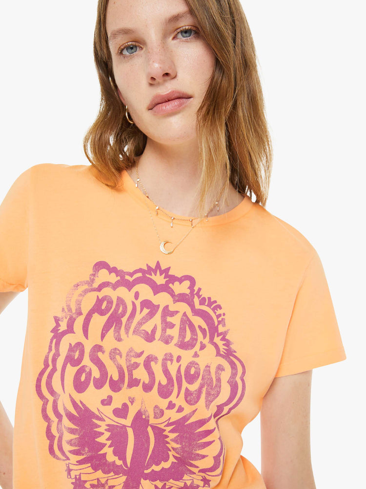 Close up view of a woman in a  faded orange tee features a purple bird graphic with text on the front.
