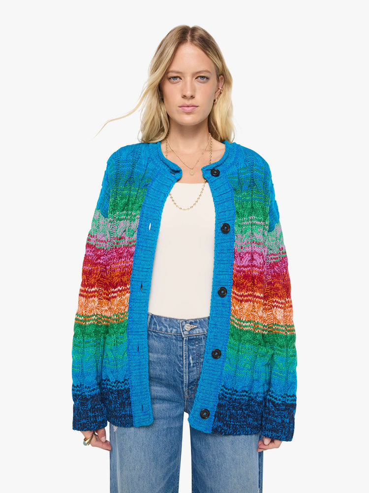 Front view of a woman in a cardigan is designed in a rainbow stripe pattern with cable knit details.