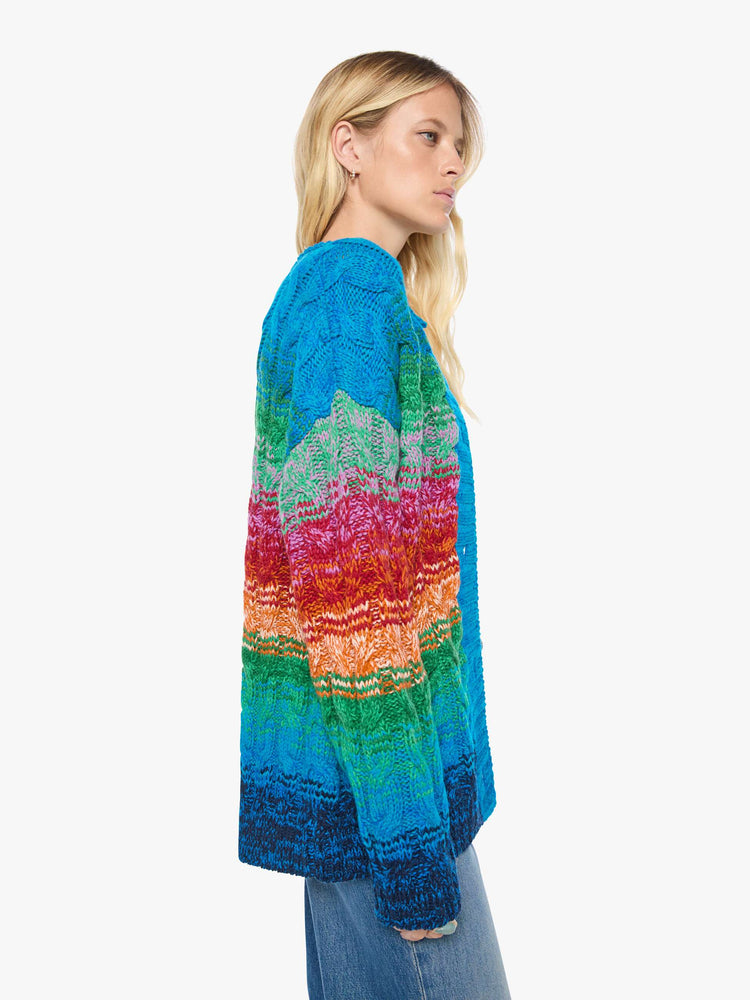 Side view of a woman in a cardigan is designed in a rainbow stripe pattern with cable knit details.