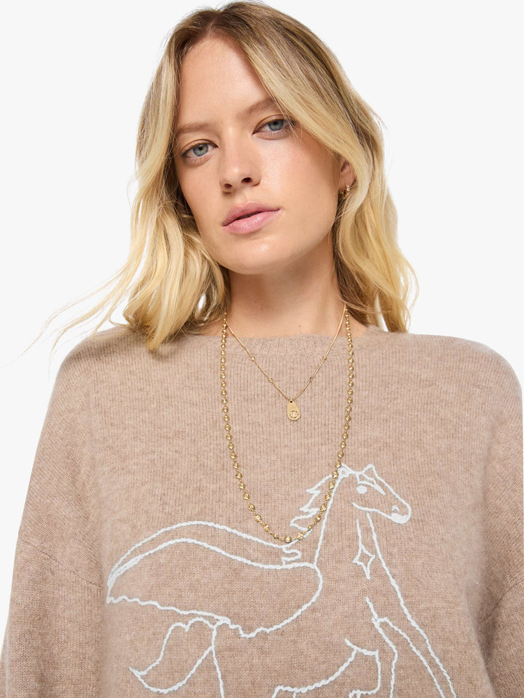 Close up view of a woman in a sweater designed in a warm tan with a knit Pegasus graphic in light blue on the front and MOTHER's name on the hip.