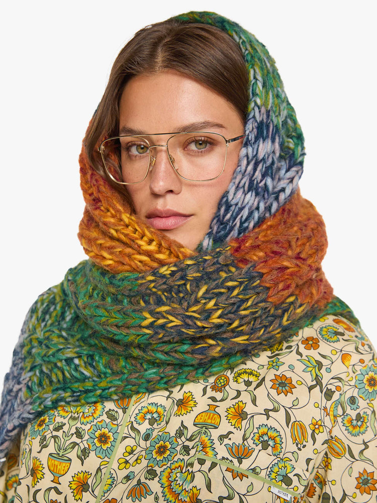 On model view of a wide, chunky scarf is made from a blend of alpaca and merino wool in a rainbow stripe pattern with cable knit details.