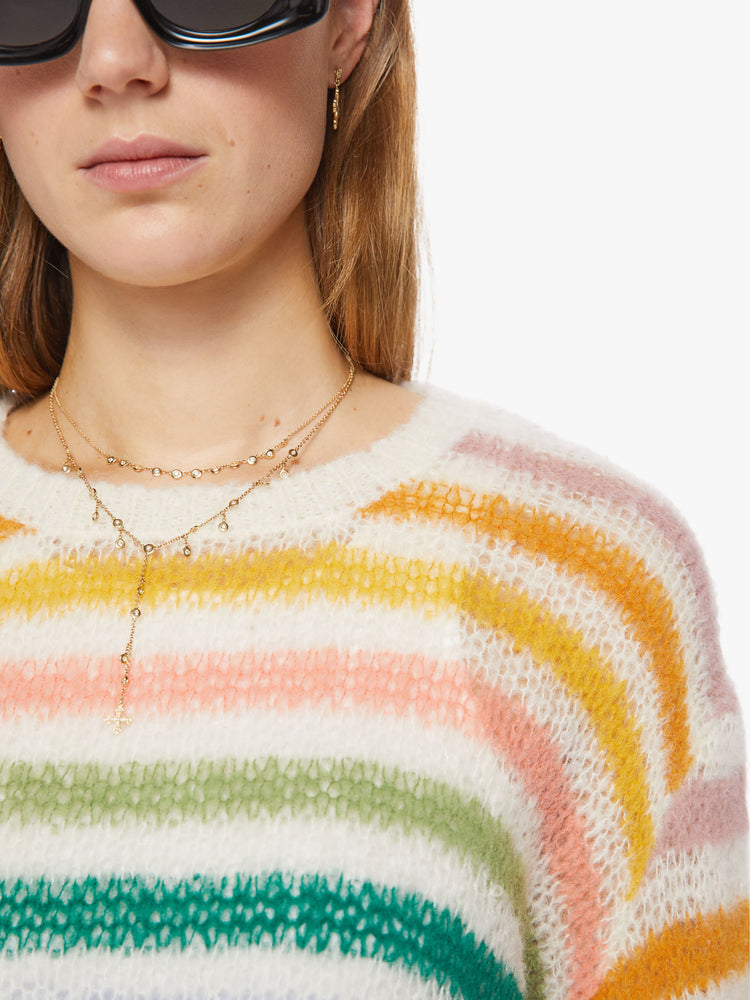Close up view of a woman oversized crewneck sweater with drop shoulders, long roomy sleeves and a hip-length hem in cream with horizontal rainbow stripes.