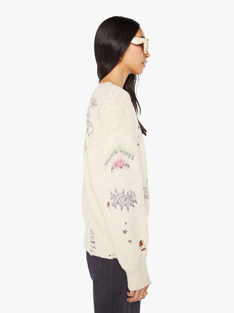 Right side view of a woman in a cream  with faded hand-drawn doodles oversized crewneck sweater with drop shoulders and ribbed hems.