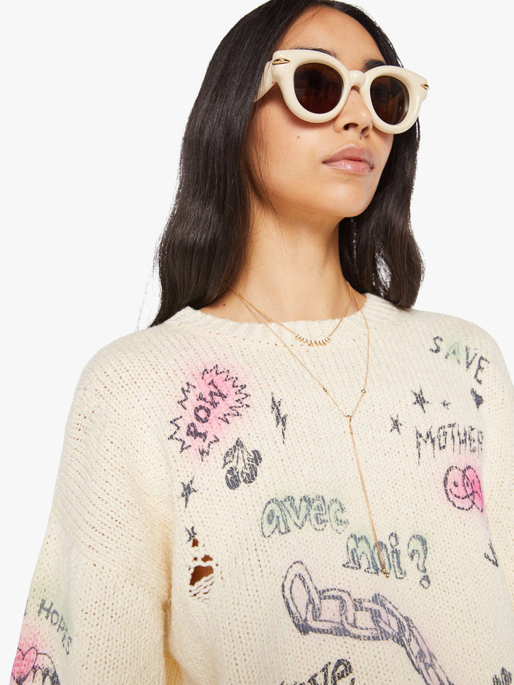 Close up view of a woman in a cream  with faded hand-drawn doodles oversized crewneck sweater with drop shoulders and ribbed hems.