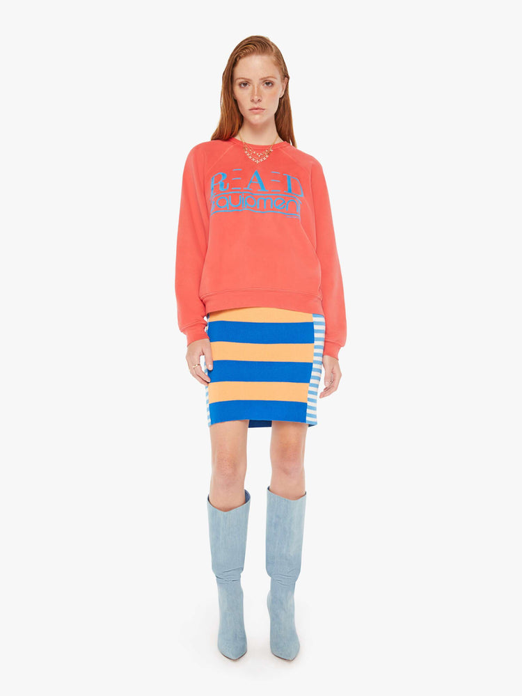A front full body view of a woman wearing a faded red sweatshirt in an oversized fit, featuring a bright blue graphic reading "RAD equipment", worn over a colorful knit dress.
