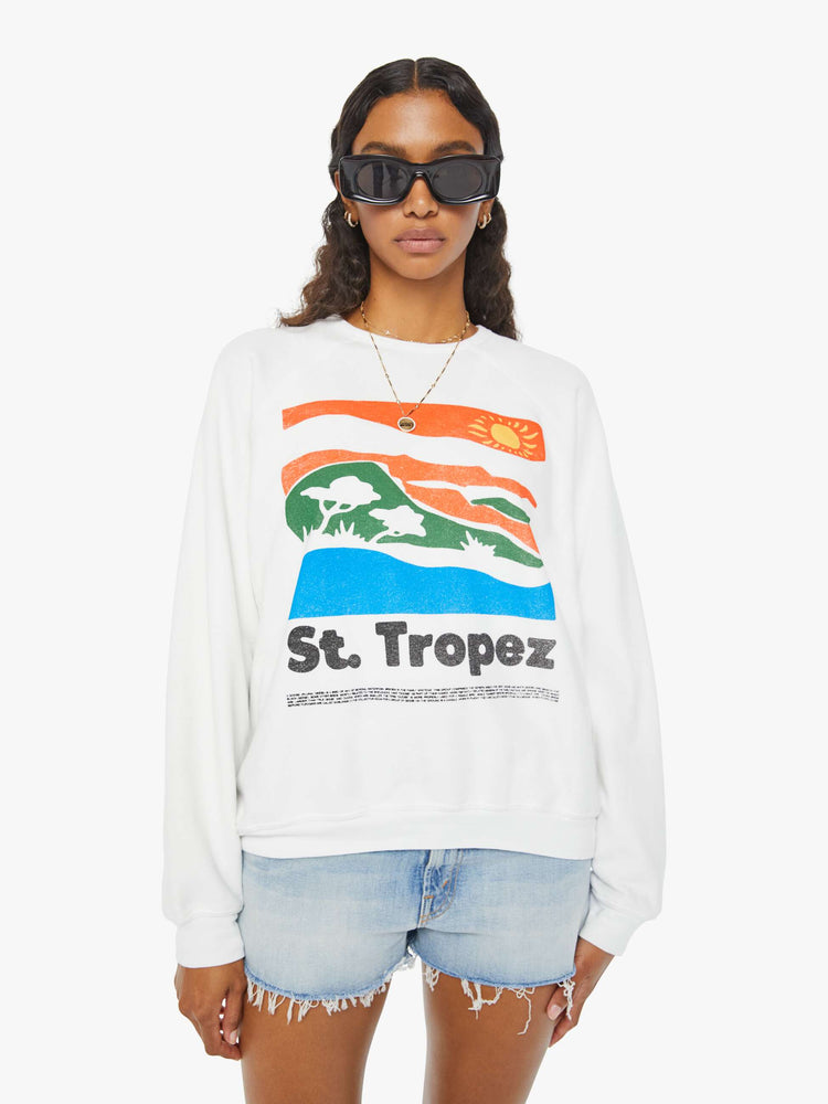 Front view of a woman in an oversized white raglan crewneck sweatshirt, with ribbed hems and a slightly cropped fit designed with a vintage-inspired St. Tropez graphic on the front.