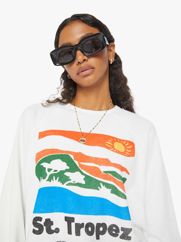Detailed view of a woman in an oversized white raglan crewneck sweatshirt, with ribbed hems and a slightly cropped fit designed with a vintage-inspired St. Tropez graphic on the front.