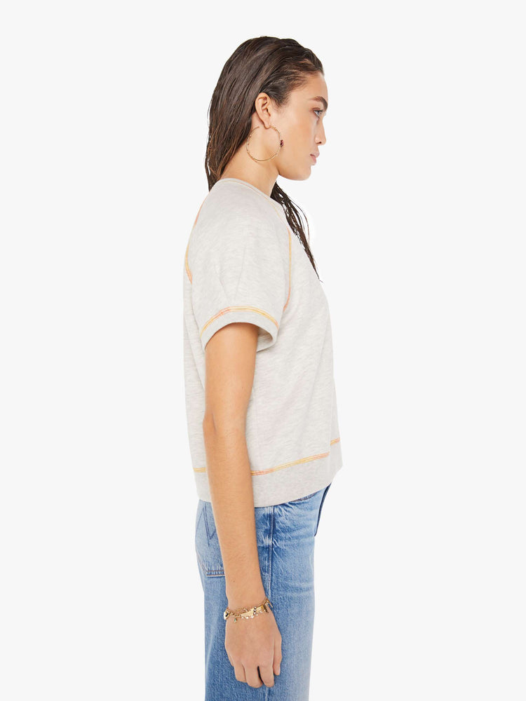 A side view of a woman wearing  a light heather grey crew neck sweatshirt featuring short raglan sleeves, a cropped body, and contrast orange thread, paired with medium blue jeans.