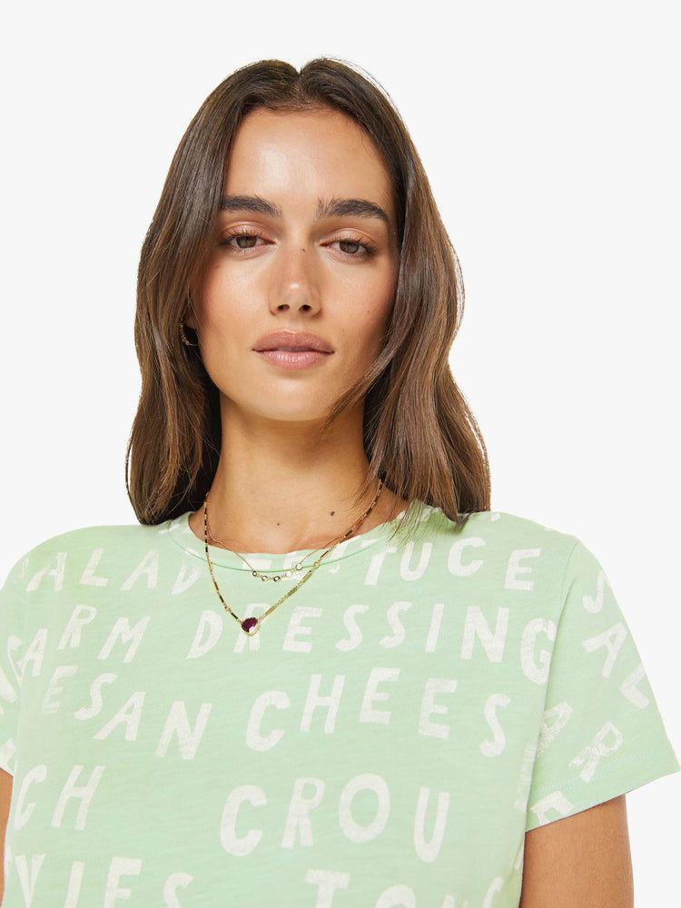 Close up view of a woman in a light green tee with a list of ingredients in faded off-white text on the front.