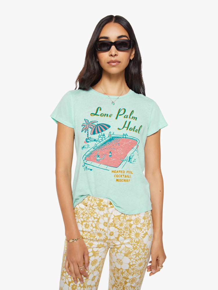 Front view of a woman in a light teal tee with a vintage-inspired hotel logo and pool scene on the front and text in teal on the back.