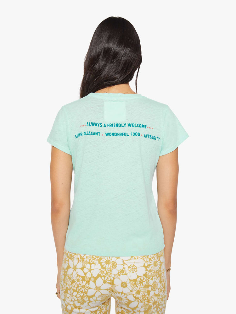 Back view of a woman in a light teal tee with a vintage-inspired hotel logo and pool scene on the front and text in teal on the back.