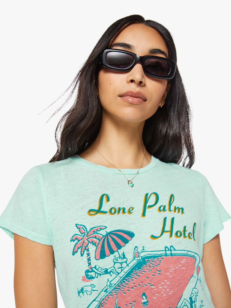 Close up view of a woman in a light teal tee with a vintage-inspired hotel logo and pool scene on the front and text in teal on the back.