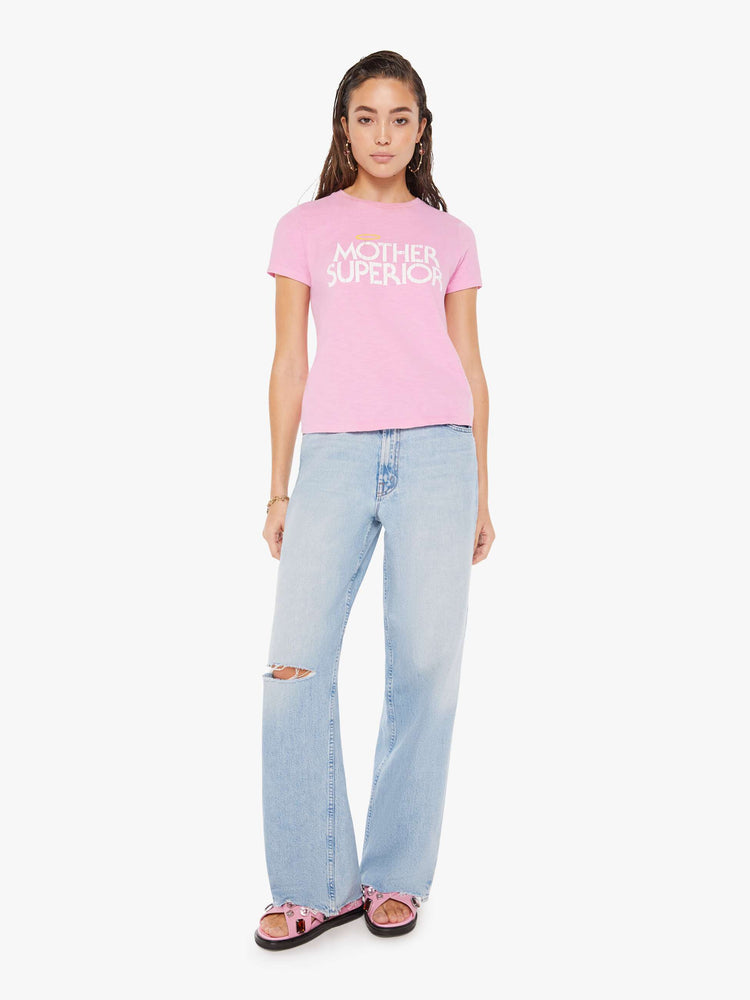 Front full body view of a woman wearing a fitted pink crew neck tee featuring a distressed graphic reading "MOTHER SUPERIOR" with an angel halo, paired with a light blue wash wide leg jean.