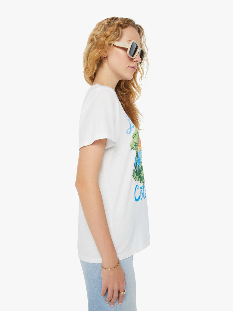 Side view of a woman in a white crewneck tee that features a hand-drawn seaside graphic with faded text in French on the front. Paired with light blue jeans. 