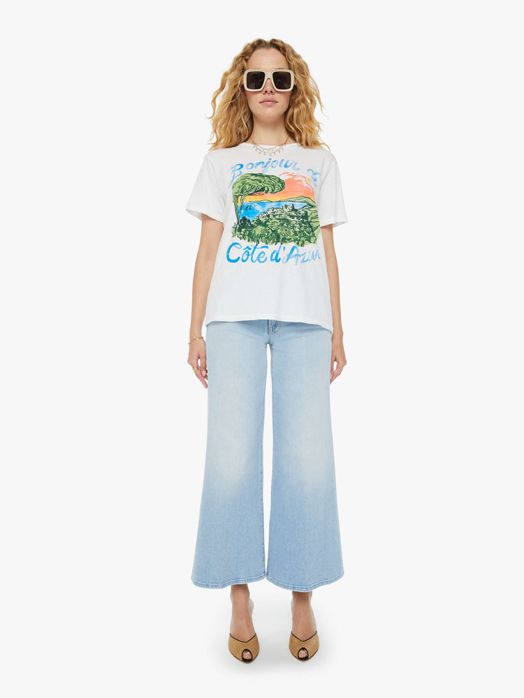 Full front view of a woman in a white crewneck tee that features a hand-drawn seaside graphic with faded text in French on the front. Paired with light blue jeans. 