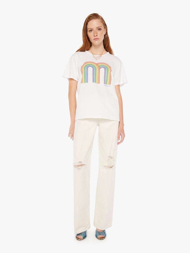 A front full body view of a woman wearing an oversized white crew neck tee featuring a large double rainbow graphic, paired with a pair of distressed wide leg jeans in off white.