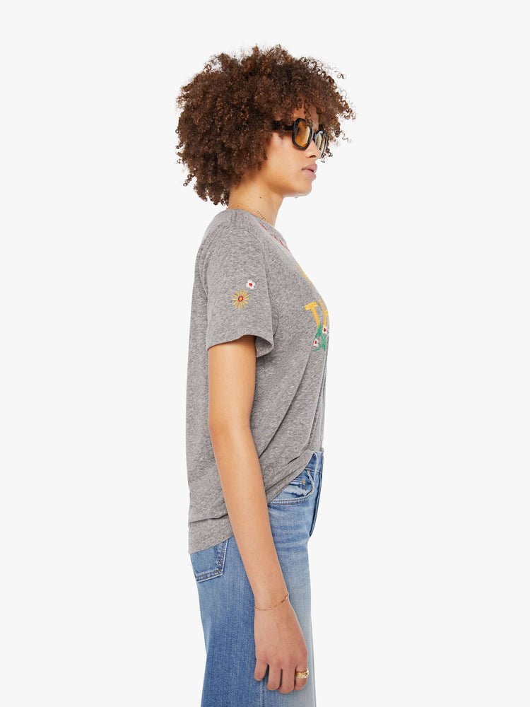 Side  view of a woman in a heathered grey tee features yellow block text on the front and colorful embroidery throughout.