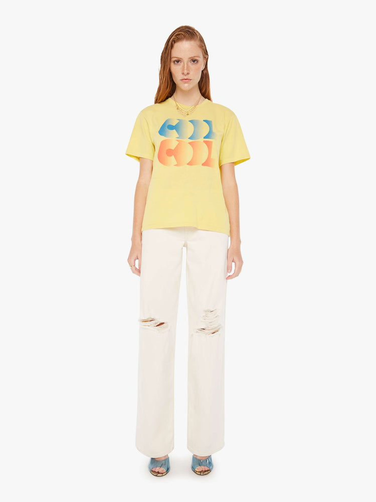 A front full body view of a woman wearing a faded yellow oversized crew neck tee featuring a red and blue graphic reading "COOL COOL", paired with off white jeans.