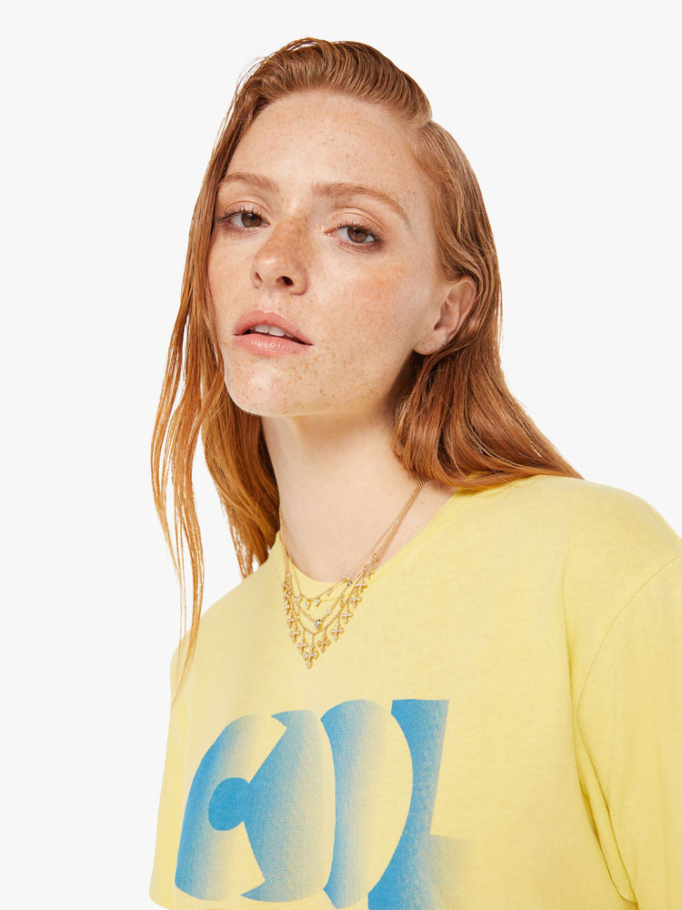 A front close up view of a woman wearing a faded yellow oversized crew neck tee featuring a blue graphic reading "COOL"