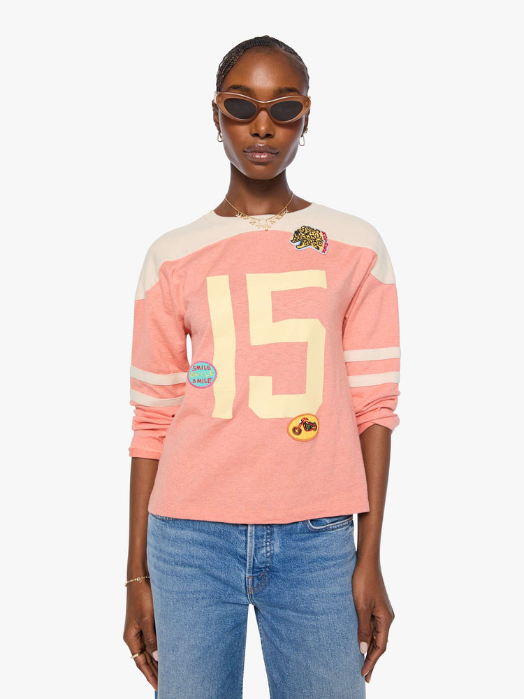 Front view of a long sleeve pale pink with an off-white panel across the shoulders, horizontal stripes on the sleeves, the number 15 in cream on the front and colorful sewn patches throughout.