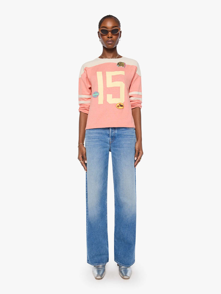 Full body view of a long sleeve pale pink with an off-white panel across the shoulders, horizontal stripes on the sleeves, the number 15 in cream on the front and colorful sewn patches throughout.