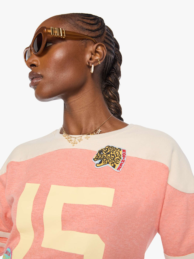 Close up view of a long sleeve pale pink with an off-white panel across the shoulders, horizontal stripes on the sleeves, the number 15 in cream on the front and colorful sewn patches throughout.