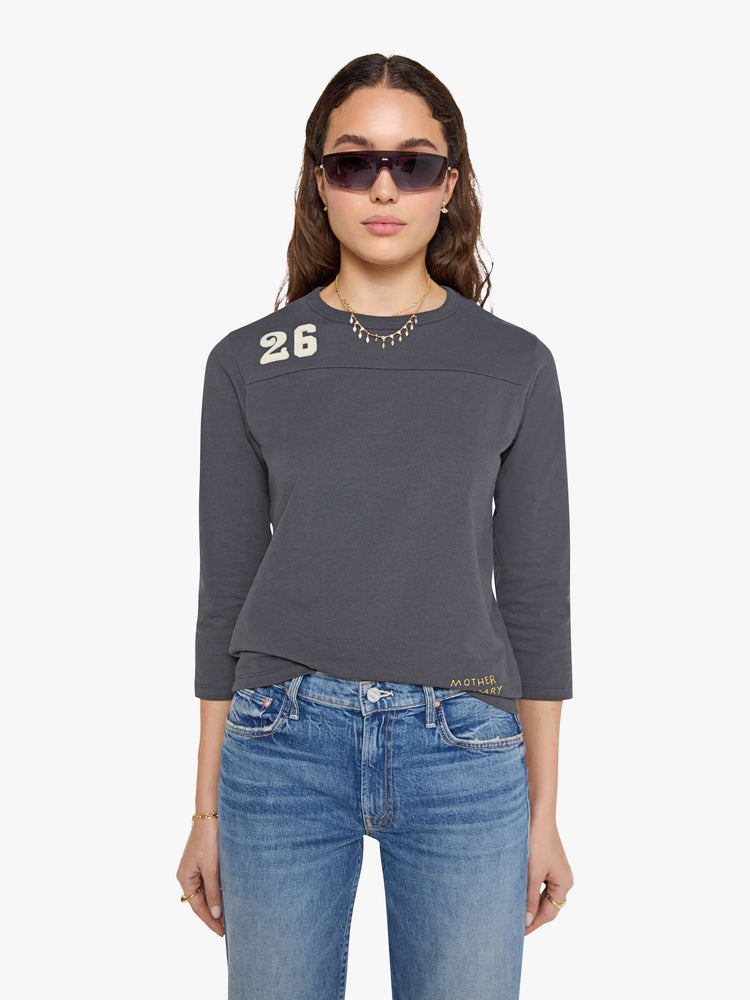 Front view of a woman in a faded black top designed with a 26 patch on the chest, ROCK across the back, and MOTHER's name embroidered in yellow at the hip.