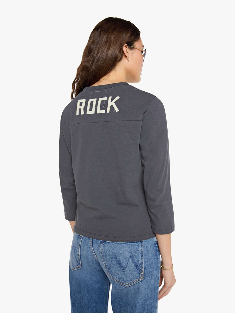 Back view of a woman in a faded black top designed with a 26 patch on the chest, ROCK across the back, and MOTHER's name embroidered in yellow at the hip.