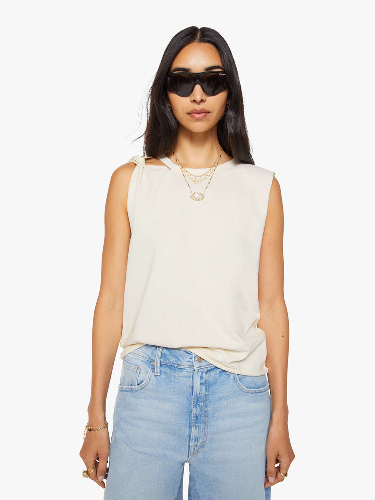 Front view of a woman in a off white sleeveless tee with a crewneck, knotted detail on one shoulder, an oversized fit and a raw hem.