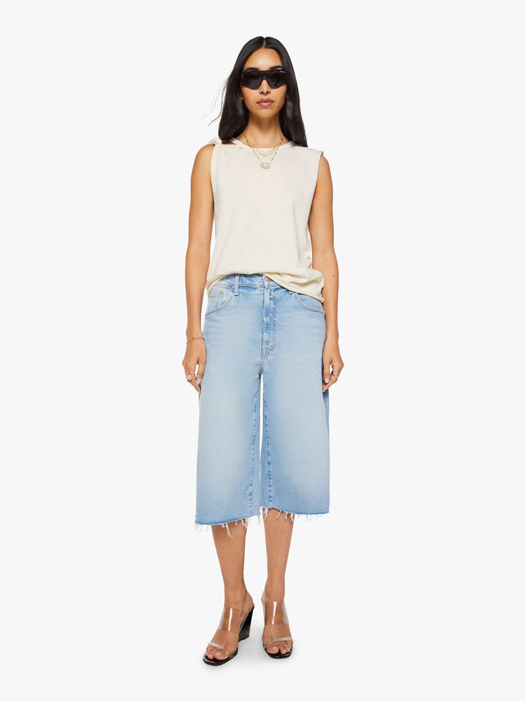 Full body view of a woman in a off white sleeveless tee with a crewneck, knotted detail on one shoulder, an oversized fit and a raw hem.
