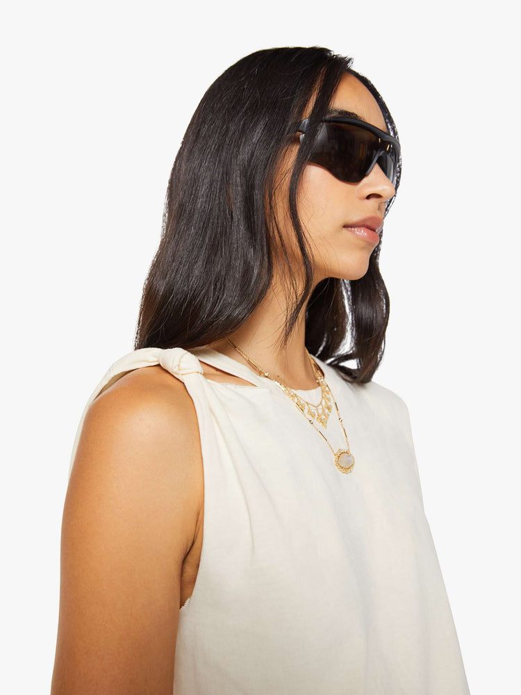 Close up view of a woman in a off white sleeveless tee with a crewneck, knotted detail on one shoulder, an oversized fit and a raw hem.