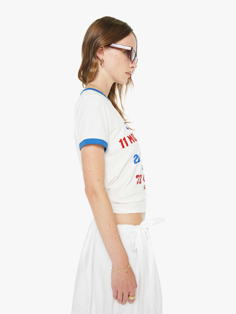 Side view of a woman in off-white with light blue trim tee and a colorful text graphic inspired by a vintage scoreboard on the front.