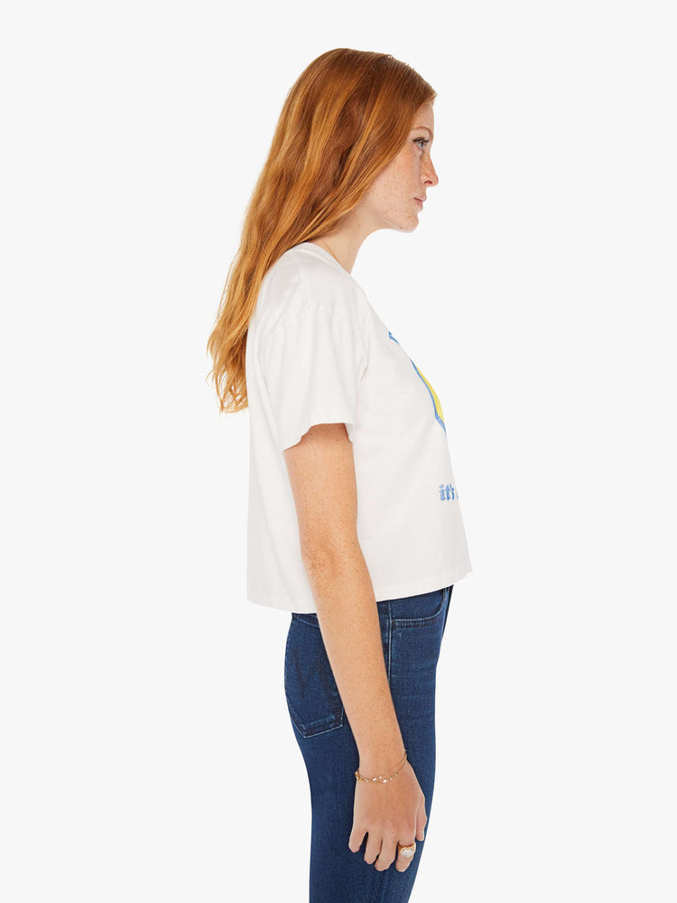 Side view of a women white oversized crewneck tee with drop shoulders, a cropped hem and a boxy fit with a faded smiley graphic with text in blue on the front.