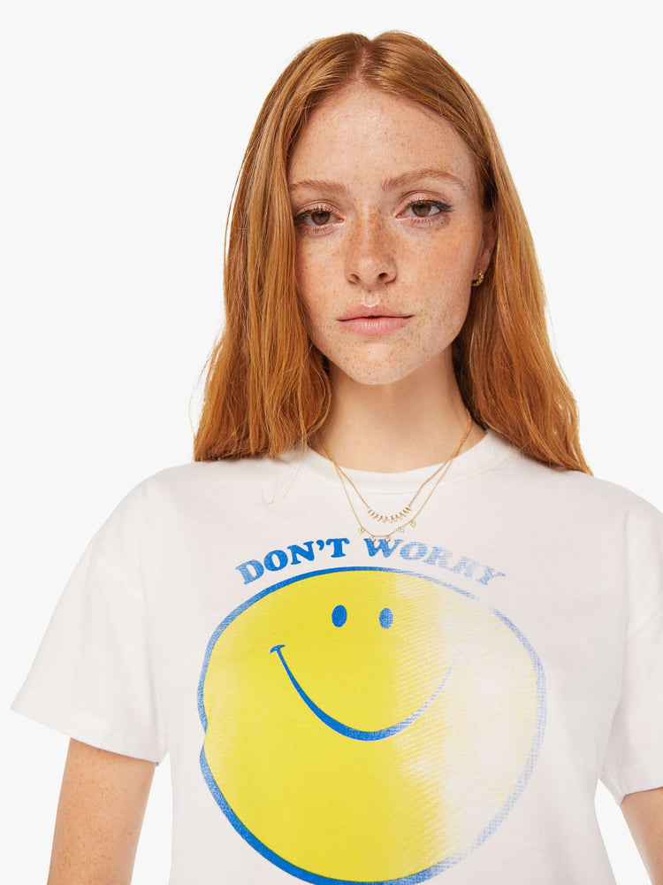 Close up view of a women white oversized crewneck tee with drop shoulders, a cropped hem and a boxy fit with a faded smiley graphic with text in blue on the front.