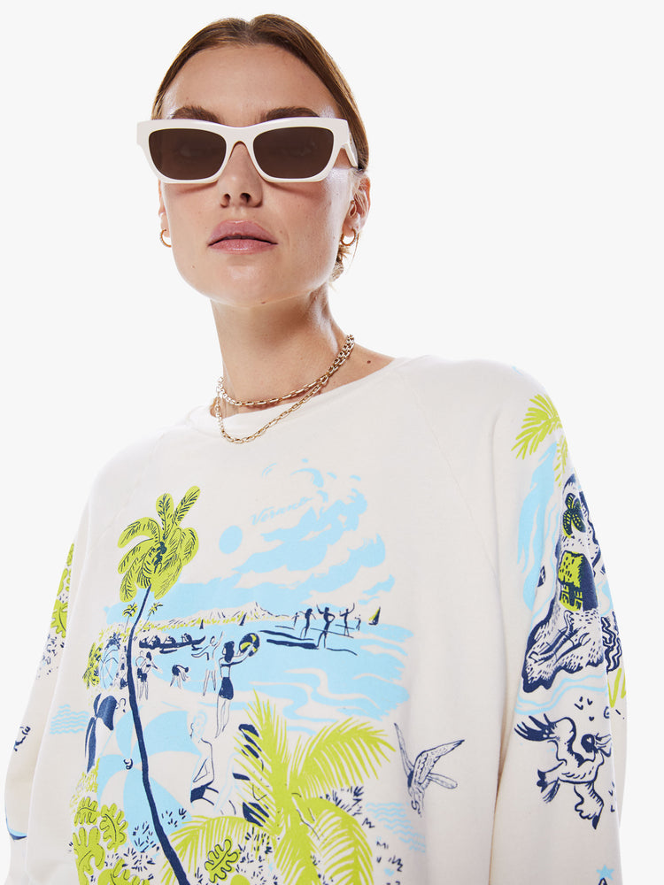 Close up view of a woman oversized crewneck sweatshirt with long balloon sleeves and a cropped cut-off hem in an off-white hue with a blue and green graphic of beachy activities and tropical plants.