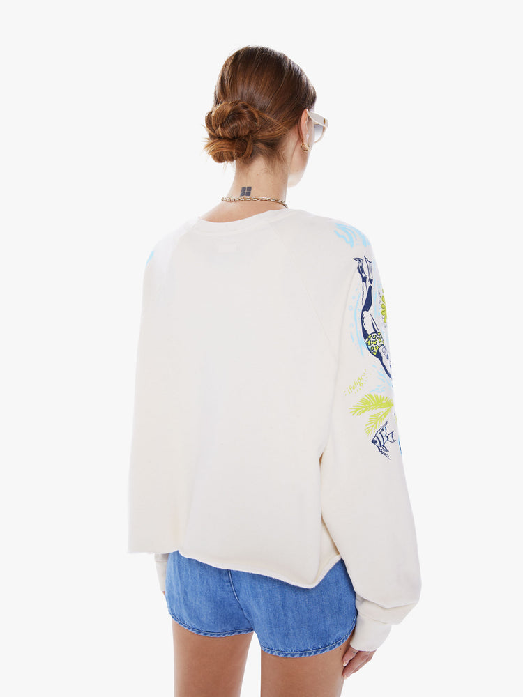 Back view of a woman oversized crewneck sweatshirt with long balloon sleeves and a cropped cut-off hem in an off-white hue with a blue and green graphic of beachy activities and tropical plants.