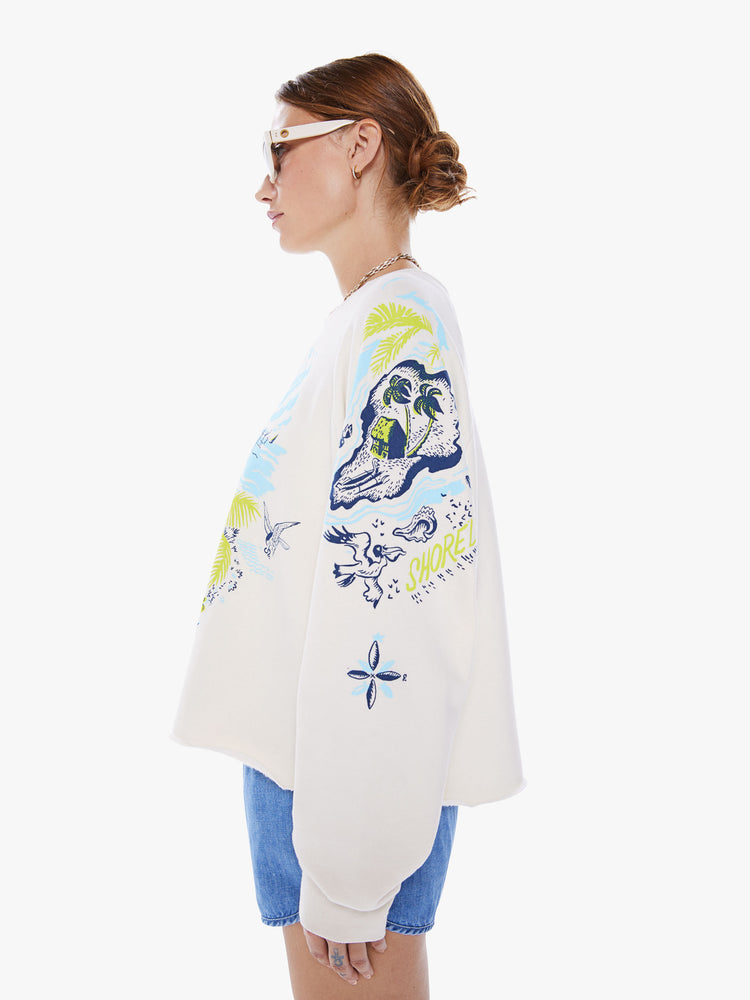 Side view of a woman oversized crewneck sweatshirt with long balloon sleeves and a cropped cut-off hem in an off-white hue with a blue and green graphic of beachy activities and tropical plants.