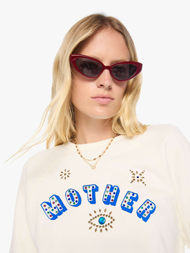 Close up view of a woman in off-white with MOTHER's name in blue block text across the chest, studded details and golden yellow stars on the back sweatshirt.